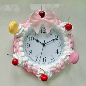 Puede incluir: Un reloj de pared decorativo con una esfera blanca y manecillas negras. El reloj está rodeado por un diseño de glaseado rosa y blanco, adornado con cerezas rojas, macarons rosas y un lazo rosa. La esfera del reloj muestra los números del 1 al 12.