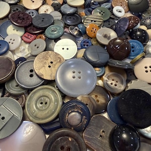 May include: A large collection of assorted sewing buttons in various shapes, sizes, and colors, including shades of gray, brown, black, blue, and cream. The buttons feature different textures, finishes, and hole counts, scattered together in a dense pile.