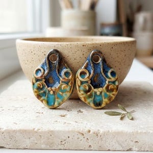 Może przedstawiać: Dwa ceramiczne wisiorki w kształcie łezki z błyszczącym szkliwem w odcieniach głębokiego błękitu, turkusu i ciepłego brązu. Każdy wisiorek ma okrągły otwór u góry i ozdobiony jest misternymi, wypukłymi wzorami w kształcie wirów.