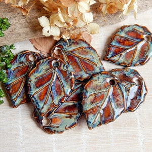 May include: A collection of handmade ceramic leaf-shaped ornaments. Each leaf features a glossy finish with a blend of brown, blue, and orange hues. The leaves have a hole at the top for hanging. Decorative items for home decor.