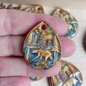 Può includere: Un ciondolo in ceramica con una glassa blu, marrone e oro. Il ciondolo presenta un design dettagliato di un orso che cammina attraverso una foresta.