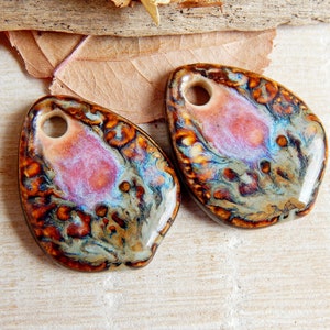 May include: Two ceramic pendants with a hole at the top. The pendants have an organic shape and are glazed with brown, orange, blue, and pink. The glaze creates a marbled effect. Each pendant is approximately 5 cm in length.