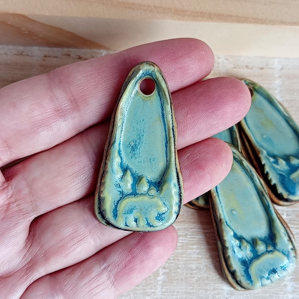 May include: A turquoise ceramic pendant with a bear and tree design. The pendant has a hole at the top for stringing. The edges are a contrasting brown color. Other similar pendants are in the background.