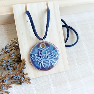 May include: A round pendant necklace featuring a white bee design on a blue and purple background. The pendant is attached to a dark blue suede cord. The necklace is displayed on a wooden stand.