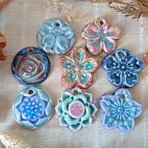 Op de afbeelding: Een verzameling handgemaakte keramische hangers in verschillende bloemen- en cirkelvormige ontwerpen. De hangers hebben een reeks kleuren, waaronder blauw, roze en turquoise, met ingewikkelde details en texturen. Elke hanger heeft een gat om te rijgen.