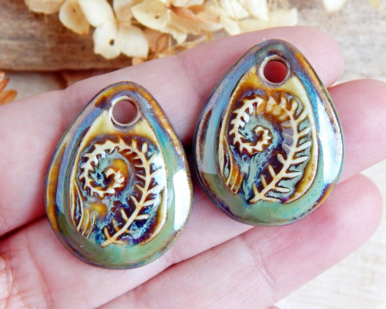 Puede incluir: Dos colgantes de cer&aacute;mica en forma de l&aacute;grima con un dise&ntilde;o de helecho. Los colgantes tienen un agujero en la parte superior para ensartar. Los colores incluyen tonos de verde, marr&oacute;n y azul, con un acabado brillante. El dise&ntilde;o del helecho es en relieve y detallado.