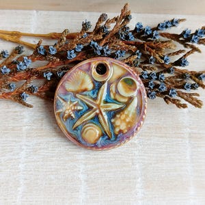 May include: A ceramic coaster with a sea life design. The coaster features a starfish, seashells, and a blue and purple colour scheme. The coaster is round and has a hole in the centre.
