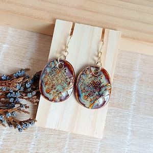 May include: A pair of handmade ceramic earrings. Each earring is an oval shape with a textured surface, featuring a blend of brown, green, and blue hues. The earrings hang from gold-colored ear wires, showcasing a unique, artistic design.