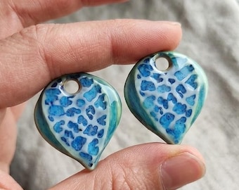 May include: Two ceramic pendants in a leaf shape, held in a hand. The pendants have a blue and white pattern with a glossy finish, and a hole at the top for stringing. The edges are a teal color.