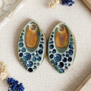 May include: Two marquise-shaped ceramic pendants featuring a glossy finish. Each piece displays a central smooth amber-toned oval surrounded by a textured pattern of raised blue and dark navy circular dots on a pale cream-colored background.