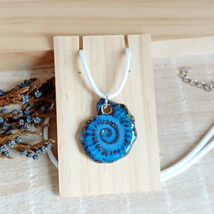 May include: A handcrafted blue ceramic pendant necklace with a spiral design. The pendant is attached to a white cord. The necklace is displayed on a wooden stand. The pendant measures approximately 1.5 inches in diameter.