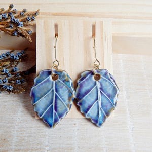 Op de afbeelding: Een paar oorbellen met blauwe en paarse, blaadjesvormige keramische hangers met gouden haakjes.