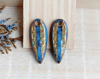 May include: Two ceramic teardrop-shaped pendants. Each pendant has a central blue stripe with textured, earthy-toned patterns. A small hole is at the top for stringing. The pendants are displayed on a light wooden surface.