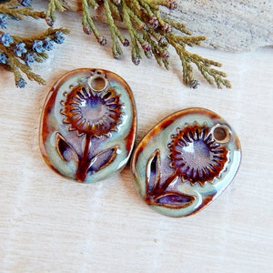 May include: Two ceramic sunflower pendants with a hole for stringing. The pendants are oval-shaped with a raised sunflower design in the centre. The colours are a mix of brown, blue, and green, with a glossy finish. Ideal for jewellery making.