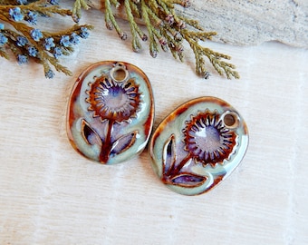 May include: Two ceramic sunflower pendants with a hole for stringing. The pendants are oval-shaped with a raised sunflower design in the center. The colors are a mix of brown, blue, and green, with a glossy finish. These pendants are ideal for jewelry making.