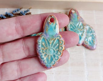 May include: A teal blue ceramic pendant with a white eye design and intricate details. The pendant has a rustic, glazed finish and a hole at the top for hanging.