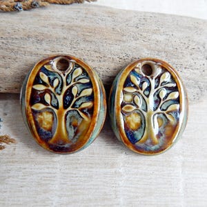 Può includere: Due ciondoli ovali in ceramica con un design ad albero della vita. I ciondoli sono marroni, blu e gialli. Ognuno ha un foro nella parte superiore per l'infilatura. Sono esposti su una superficie di legno chiaro. Misurano circa 3 cm.