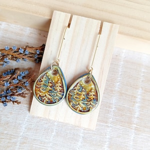 May include: Teardrop-shaped earrings with a forest scene. The earrings feature a gold-colored metal hook and a detailed ceramic pendant with a blue border. The forest scene includes trees in shades of brown, green, and yellow.