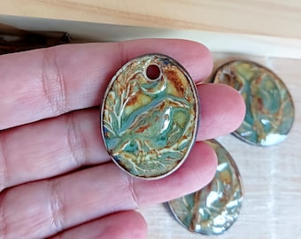 May include: Oval-shaped ceramic pendant with a nature-inspired design. The pendant features a landscape scene with shades of green, brown, and blue, framed by a silver-colored metal edge. A small hole is at the top for stringing.