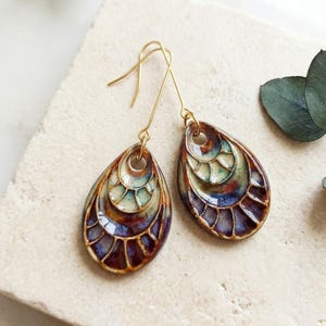 May include: A pair of teardrop-shaped earrings featuring a multi-layered ceramic design with a glossy finish. The colors transition from a deep purple and brown at the base to a lighter cream and teal toward the top, suspended by gold-toned hooks.