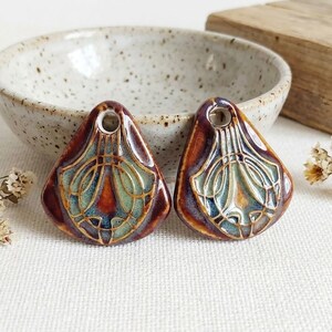 May include: Two teardrop-shaped ceramic pendants featuring a glossy glaze in shades of deep brown, teal, and amber. Each piece has a raised, intricate geometric pattern with a central hole at the top for hanging, showcasing a smooth finish.