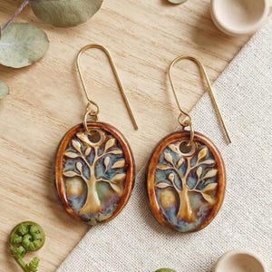 Può includere: Un paio di orecchini pendenti di forma ovale con un motivo ad albero in rilievo color crema su uno sfondo multicolore blu, verde e marrone. Gli orecchini sono sospesi a ganci in metallo dorato e presentano una finitura lucida e smaltata.