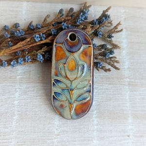 Könnte beinhalten: Ein Keramik-Anhänger mit einem floralen Design in Blautönen, Grün- und Brauntönen. Der Anhänger ist oval geformt und hat oben ein Loch zum Aufhängen.