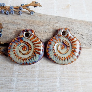 May include: Two ceramic pendants with a spiral design in shades of orange, brown, and blue. Each pendant has a hole at the top for stringing. The pendants are displayed on a piece of driftwood with blue floral accents.