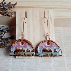Peut inclure: Une paire de boucles d'oreilles en céramique en forme de demi-cercle avec un fil de cuivre. Les boucles d'oreilles sont décorées d'une scène de paysage dans des tons de brun, violet et blanc.