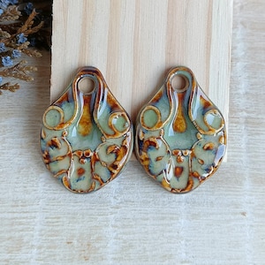 Può includere: Due ciondoli in ceramica con una glassa verde e marrone. I ciondoli hanno un design floreale dettagliato e un foro nella parte superiore per l'appeso.