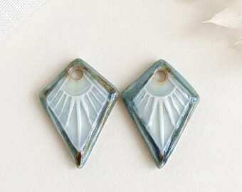 Puede incluir: Dos colgantes de cerámica en forma de diamante con un diseño de rayos de sol. Los colgantes son de color azul claro con detalles blancos y un borde marrón. Cada uno tiene un pequeño agujero en la parte superior para ensartar.