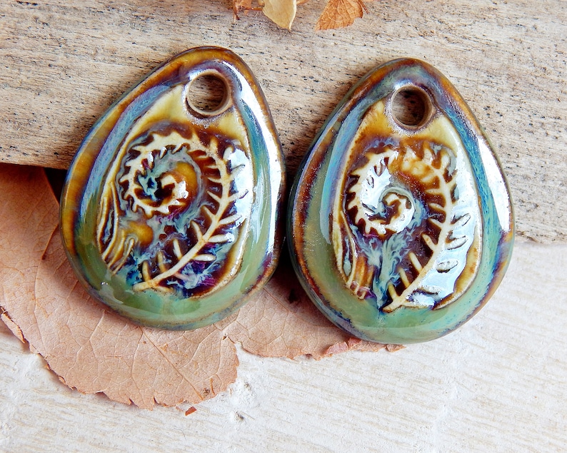 Puede incluir: Dos colgantes de cer&aacute;mica en forma de l&aacute;grima con un dise&ntilde;o de helecho. Los colgantes tienen un agujero en la parte superior para ensartar. La combinaci&oacute;n de colores incluye tonos de verde, marr&oacute;n y azul, con un acabado brillante. El dise&ntilde;o del helecho es en relieve.