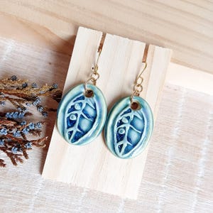 Puede incluir: Un par de pendientes ovalados con un diseño de hoja. Los pendientes son de color azul claro con un patrón de hoja azul más oscuro y ganchos dorados. Los pendientes se exhiben en un soporte de madera.