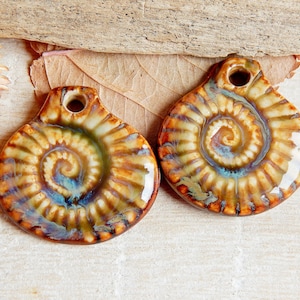 Puede incluir: Dos colgantes de cerámica con un diseño en espiral en tonos marrones, verdes y azules. Los colgantes tienen un agujero en la parte superior para colgar.