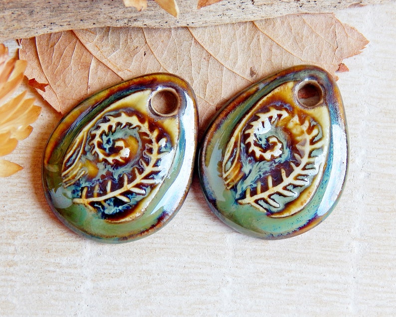 Puede incluir: Dos colgantes de cer&aacute;mica con forma de l&aacute;grima. Cada colgante presenta un dise&ntilde;o de helecho en relieve en crema sobre un fondo esmaltado verde, marr&oacute;n y azul. Un peque&ntilde;o agujero en la parte superior para ensartar. Suministros para la fabricaci&oacute;n de joyas.