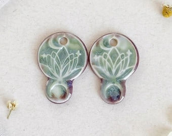 May include: Two ceramic pendants with a green and white lotus flower design. Each pendant has a hole at the top and a crescent moon and star design. The bottom of each pendant has a purple and green color.