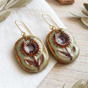 Puede incluir: Un par de pendientes colgantes de cerámica de forma ovalada con un diseño de girasol pintado a mano en tonos marrón y verde salvia. Los pendientes cuelgan de ganchos de metal dorado y tienen un acabado brillante y suave en la superficie.
