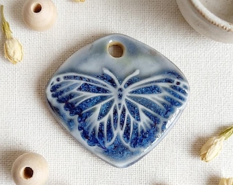 May include: A diamond-shaped ceramic pendant featuring a glossy finish with a deep blue and white butterfly motif. The pendant has a single hole at the top for suspension and is surrounded by several small wooden beads and dried flower buds.