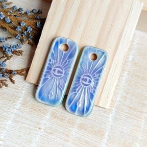 May include: Two rectangular, blue and white ceramic pendants with a sunburst and eye design. Each pendant has a hole at the top. The pendants are on a light wood surface.