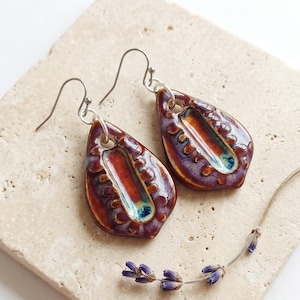 Peut inclure: Une paire de boucles d'oreilles en céramique en forme de goutte avec un vernis brillant dans des tons de violet profond, orange brûlé et des touches de bleu sarcelle. Elles présentent un bord texturé en relief autour d'un centre lisse.