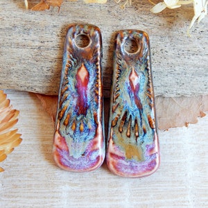 May include: Two ceramic pendants with a hole at the top for hanging. The pendants are rectangular with a flared bottom. The glaze has a mix of colors including brown, red, blue, and gold. The pendants have a unique, artistic design.