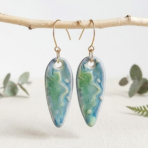 Può includere: Un paio di orecchini in ceramica a forma di goccia con una smaltatura lucida nei toni del verde schiuma di mare, azzurro cielo e bianco. Gli orecchini pendono da sottili ganci in metallo dorato, con una finitura liscia e marmorizzata.