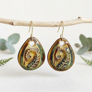 May include: A pair of teardrop-shaped ceramic earrings featuring a glossy glaze in shades of brown, green, and cream. Each earring displays an embossed fern leaf pattern and is suspended from a gold-toned metal hook for pierced ears.