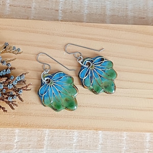Peut inclure: Une paire de boucles d'oreilles en forme de feuille avec un motif émaillé bleu et vert. Les boucles d'oreilles ont des crochets d'oreilles argentés et un motif veiné détaillé. Les boucles d'oreilles sont posées sur une surface en bois brun clair.