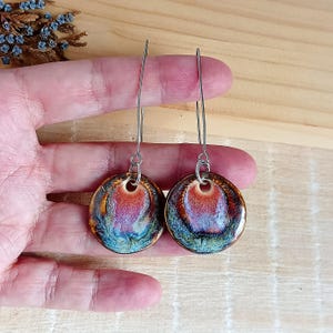 Könnte beinhalten: Ein Paar handgefertigte Keramikohrringe mit einem einzigartigen, farbenfrohen Design. Die Ohrringe haben eine runde Form mit einer lebendigen Mischung aus Rot-, Lila- und Blautönen. Sie hängen an langen, silberfarbenen Ohrdrähten und sind ein auffälliges Accessoire.