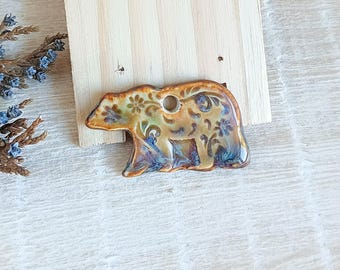 May include: A ceramic bear pendant with a hole for hanging. The bear is tan with floral and swirl designs in blue, green, and brown. The pendant is on a light wood background.