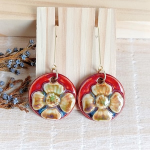 Puede incluir: Un par de pendientes de cerámica hechos a mano. Cada pendiente presenta una base circular roja con un diseño floral en tonos crema, verde y azul. Los pendientes cuelgan de alambres dorados.