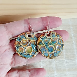 Peut inclure: Une paire de boucles d'oreilles en céramique faites à la main. Chaque boucle d'oreille est un disque rond avec un motif texturé en forme de bulles dans des tons de bleu, marron et vert. Les boucles d'oreilles sont suspendues à des crochets argentés.