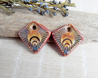 May include: Two ceramic square-shaped pendants with a hole at the top. The pendants are red with a brown and white sunburst design.