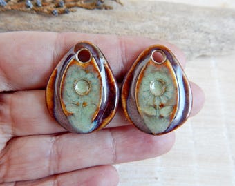 May include: Two brown ceramic teardrop-shaped pendants with a green and white floral design. The pendants have a hole at the top for hanging.